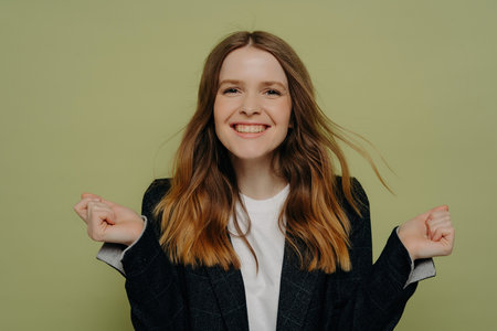 Achievement! Smiling young woman holding hands up, demonstrating excitement, looking at camera, wearing dark jacket and white top. Posing against a light studio background. human emotions concept.の写真素材