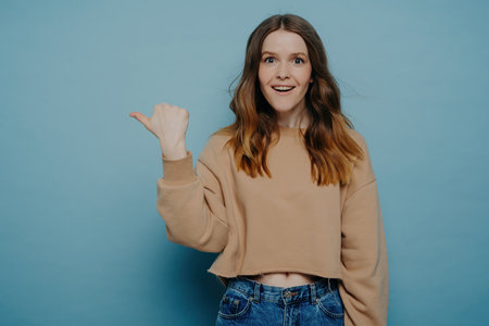 Amused pretty young female with ombre hair, in casual light brown sweater and jeans, pointing finger at something to attract attention. Standing against a blue wall. Advertising and promotion.の写真素材