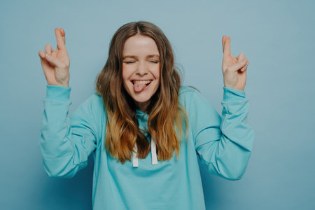 Happy crazy teenage girl with wavy ombre hair, eyes closed, fingers crossed, sticking tongue out, standing against blue wall in studio, carefree and having fun.の写真素材