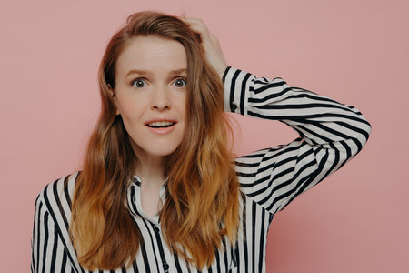 Attractive young woman in stripy black and white blouse, touching head in astonishment, expressing surprise against pink studio wall. Emotion concept.の写真素材