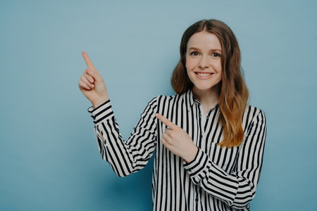 Cute teenage girl in striped shirt, shining smile, pointing up with forefingers at copy space for advertisement. Positive female advertising product, standing isolated on blue background.の写真素材