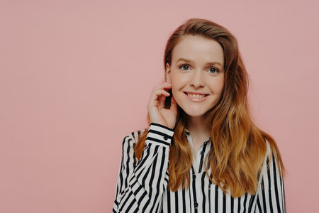 Smiling young woman in stripy blouse, wavy ginger hair, touching face, posing over light pink studio background.の写真素材