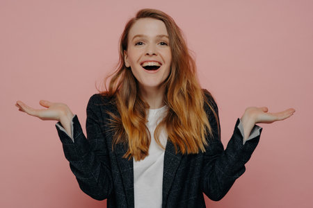 Laughing young woman in dark formal jacket and white top, raising hands with open palm gesture, looking amused and surprised, posing against pink wall.の写真素材
