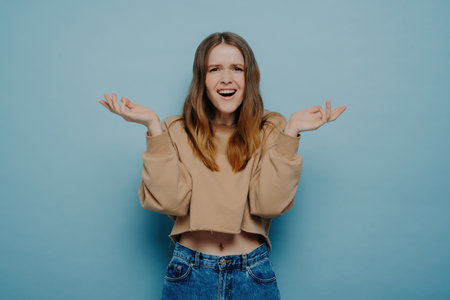 Studio shot of attractive amazed teenage girl in stylish casual outfit, cannot believe her eyes, shrugging shoulders with a surprised face expression, looking at the camera, isolated on a blue background.の写真素材