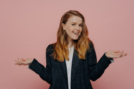 Happy young woman with medium length ginger hair, wearing dark formal jacket and white top, raising hands to sides, indoor isolated on pink background. body language concept.の写真素材