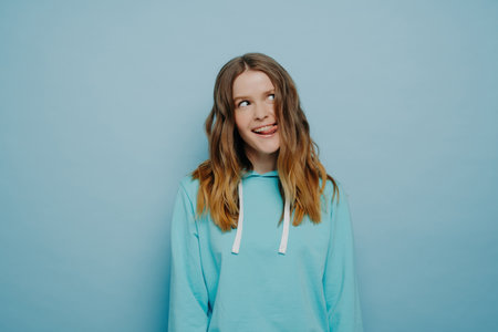 Carefree pretty teenage girl, wavy light hair, casual blue hoodie, making funny expression, showing tongue, alone on blue background.の写真素材