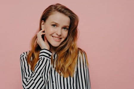 Beautiful young woman in stripy black and white blouse, wavy ginger hair, touching face, posing on light pink studio background.の写真素材