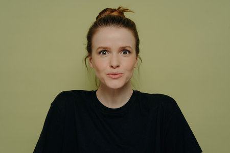 Wide-eyed woman in black tee, hair in bun, expressing surprise, staring at camera on green backdrop.の写真素材