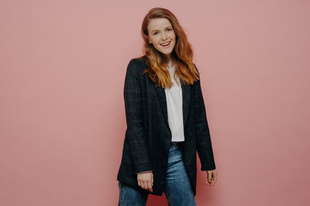 Stylish young woman in plaid blazer and jeans, posing carefree and expressing positive emotions in studio, isolated on pink wall background. happy people concept.の写真素材