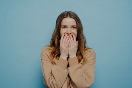 Amazed European teenage girl in casual clothes, laughing and covering mouth with hands, expressing positive emotions while posing against a blue wall in a studio.の写真素材