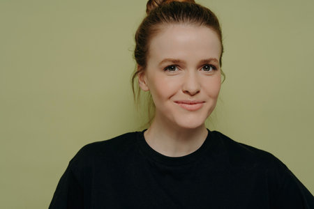 Close-up of cute female student in black tee, hair in bun, smirking and trying not to laugh, isolated on light green backdrop.の写真素材