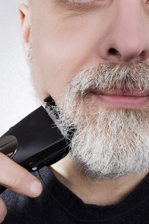 Gray-haired man shaves his beard with an electric razorの写真素材