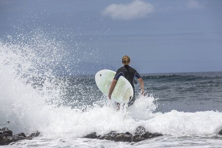 The surf enters the water. Male surfer entering the sea with his board in a black surfing suit. Tenerife, Spain.の写真素材