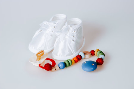 A pair of white baby shoes and a pacifier are on a white surface. The pacifier is colorful and has a blue and red designの写真素材