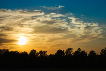 Landscape scene of golden sunset behind smooth clouds floating over blue sky with silhouette trees. Beautiful nature yellow evening at countryside, powerful summer sunlight. Dramatic scenic, contrast.の写真素材