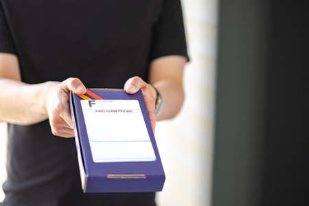 Messenger carry box package with first class label at front door, depth of field. Man in black clothes deliver priority packet at customerâs house, blurred background. Express delivery parcel conceptの写真素材