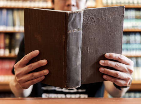 Man reading law dictionary with blurred bookshelf background, depth of field. Student with opened ancient big brown hardcover book in library. Knowledge, education concept. Clever people lifestyle.の写真素材