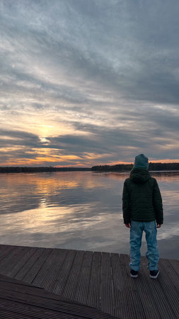 A boy stands on a pier and looks at the sunset over the lakeの写真素材
