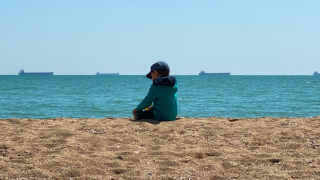 boy sitting on the beach and watching the cargo ship in the seaの写真素材