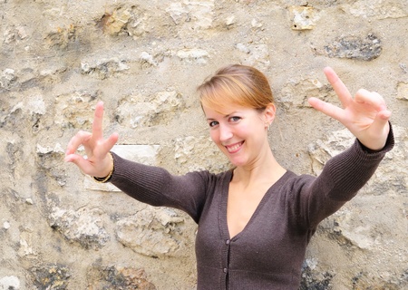 portrait of young blond smiling woman saying  V  as Victory with her two hands against ancient wall in the street の写真素材