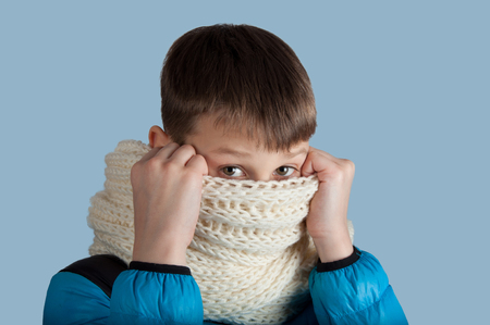 Cute boy in blue jacket and white scarf hide nose isolated on blue backgroundの写真素材