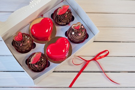 confectionery. Brown cupcakes, red glazed mousse hearts and small honey cakes in box lay on white wooden table near red bow knot. Focus on red cakesの写真素材