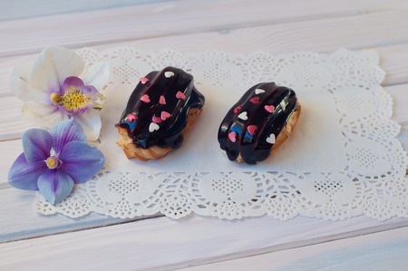 Two eclairs with dark chocolate cover and small confectionery heart sprinkles lay on white lace serviette on wooden table near orchid flowersの写真素材