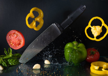 Kitchen knife and bright vegetable pieces food levitation on dark black background with water splashes. Fresh red tomato, green, yellow pepper, garlic, basil leaves, pepper seeds. Restaurant menu, recipe concept. High quality photoの写真素材