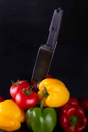 Kitchen knife standing in pile of bright vegetables on black background. Fresh red tomato, green, yellow pepper. Restaurant menu, recipe concept. Vertical photo.の写真素材