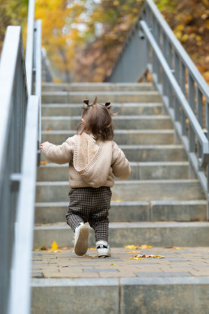 Toddler in brown pants and beige sweatshirt stepping on stairs in autumn park. Rear view. Happy childhood, walking outdoors. High quality photoの写真素材