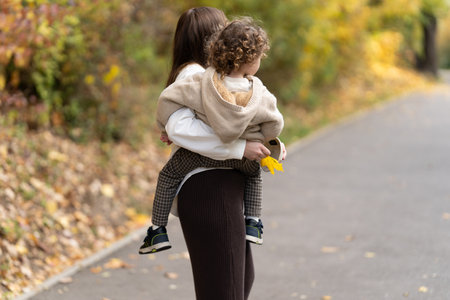 Young mom with brown hair holding small son, toddler on her hands. KId and his mother standing on autumn park background, rear view. Happy maternity concept. High quality photoの写真素材