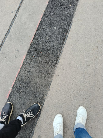 Vertical photo of old light grey and new dark asphalt. White and black human shoes standing on...