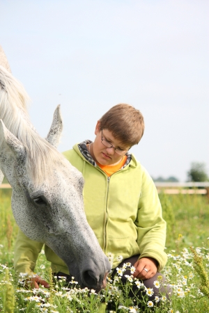 Happy teenager boy and white horse at the field with flowersの写真素材