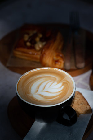 A cup of hot latte art coffee served with biscuit and pastry on wooden table in cozy cafe atmosphere with natural morning light.の写真素材