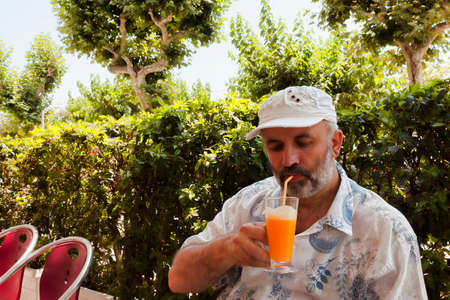 A mature man with a gray beard in a baseball cap sits in a shady cafe and drinks orange juiceの写真素材