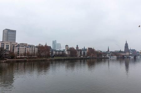 FRANKFURT (MAIN), GERMANY - DECEMBER 2, 2024: A panoramic view of the Frankfurt am Main skyline, featuring the Main River, historic buildings including the Kaiserdom (Frankfurt Cathedral), modern skyscrapers, and a grey, overcast skyの写真素材