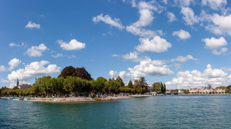 A sweeping panoramic view of the serene Lake Constance (Bodensee) in Konstanz, Germany, under a brilliant blue sky dotted with scattered white clouds. The calm, turquoise waters stretch across the foregroundの写真素材