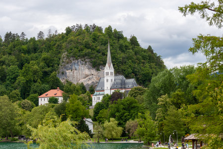 A picturesque view across the serene waters of Lake Bled, showcasing the charming town of Bled nestled at the foot of lush green hills. Prominently featured are the steeple of the Church of St. Martinの写真素材