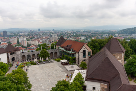 An expansive elevated view looks down upon the courtyard and buildings of Ljubljana Castle, with the sprawling city of Ljubljana and distant mountains visible under a cloudy skyの写真素材