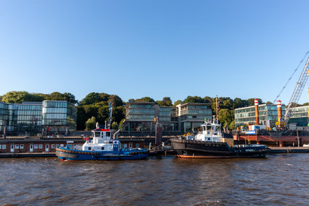 HAMBURG, GERMANY - AUGUST 12, 2024: Tugboats moored on the Elbe River in Hamburg, Germany. Scenic view of the harbor with modern office buildings and blue skyのeditorial素材
