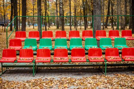 Empty sports grandstand in the leaves in the fall. Yellow leaves lie on a seat of a tribune. Plastic seats required are covered with tree foliage in the fall.の写真素材