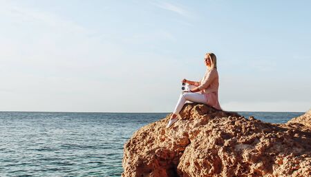 Blonde girl in sunglasses sits on a stone near the sea. A woman in pink clothes and sunglasses looks at the sea, enjoys the rest on a trip.の写真素材