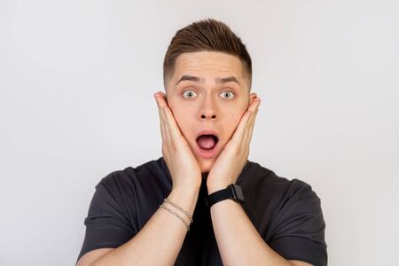 Shocked young man holding his hands on his cheeks, looking with surprised eyes and opened his mouth...