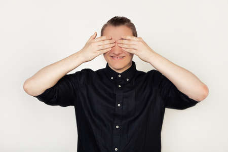 Handsome young guy student in a black shirt, smiling, closed his eyes with his hands so as not to...