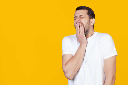 A modern young man with a beard in a white T-shirt yawns boredly against a yellow background, tired of covering his mouth with his hand. Restlessness and sleepinessの写真素材