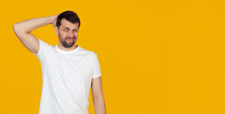 Young man with a beard in a white t-shirt worried about a problem, with a hand on his head, nervous and anxious about the crisis, stands on an isolated yellow background.の写真素材