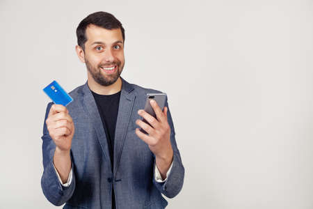 Young businessman man with a beard in a jacket, Handsome man using a credit card to pay online using a smartphone. Portrait of a man on a gray background.の写真素材