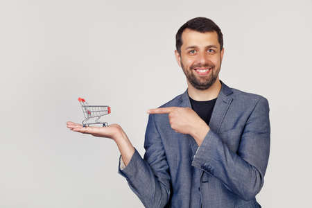 Young businessman man with a beard in a jacket looks at the camera and points to the shopping cart on his hand, showing with his hand and pointing with his fingerの写真素材