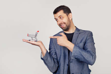 Young businessman man with a beard in a jacket looks and points to a shopping cart on his hand,...