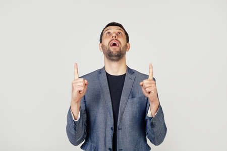 Young businessman with a smile man with a beard in a jacket, amazed and surprised, looking up and pointing with his fingers and raised hands. Portrait of a man on a gray background.の写真素材